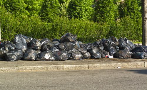 A long row of black plastic rubbish bags filled with waste materials is piled along the curb of a paved street, with a concrete edge separating the bags from the road. The bags appear to be tightly packed and are stacked in multiple layers, stretching across the foreground of the image. Behind the rubbish bags, a lush line of green trees and bushes provides a natural backdrop, indicating an outdoor urban or suburban environment. The scene is illuminated by daylight, with sunlight casting subtle shadows on the bags and pavement. This image depicts an accumulation of collected waste awaiting removal or disposal, relating to professional rubbish collection services such as those offered by Commercial Waste Mill Hill in the local North London area, close to Mill Hill postal district.