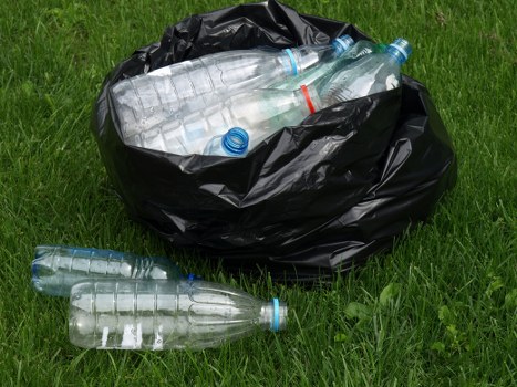 A black plastic rubbish bag sits on a grassy surface outdoors, filled with clear plastic water bottles, some with blue caps and one with a red band, which are partially visible through the bag's material. Several additional empty plastic bottles are scattered on the grass nearby, displaying transparent, smooth textures with visible ridges on their bodies and caps. The setting appears to be an outdoor area, possibly a garden or verge, with vibrant green grass surrounding the waste, and the natural daylight illuminating the scene evenly. This image exemplifies typical rubbish collection and disposal activities undertaken by Commercial Waste Mill Hill in the suburban area, including the handling of recyclable plastic bottles as part of general rubbish removal services in the Mill Hill locale. The scene reflects proper waste containment practices for curbside or outdoor collection, with a focus on environmental responsibility and efficiency in rubbish management.