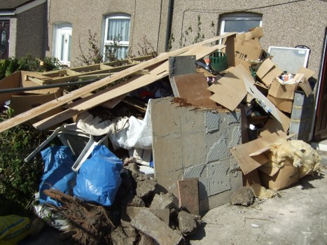 A large pile of mixed refuse and construction debris situated on a pavement outside a residential building with pebbledash exterior walls and small front gardens. The pile includes broken concrete blocks, wooden planks, cardboard boxes, and fabric waste, with some cardboard being flattened and others still intact. The debris appears messy, with materials overlapping and scattered. In the background, part of the building's windows and small garden areas with shrubs are visible. The scene is illuminated by daylight, emphasizing the textures and colors of the various materials, which include grey concrete, brown wood, and the brown and white tones of cardboard and fabric waste. This image exemplifies typical rubbish awaiting removal service, possibly handled by companies like Commercial Waste Mill Hill, serving the Mill Hill area for waste clearance solutions.