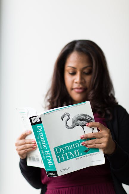 A woman with medium skin tone and long, dark wavy hair is holding an open book titled 'Dynamic HTML,' which features a black and white illustration of a flamingo on the cover. She is dressed in a dark